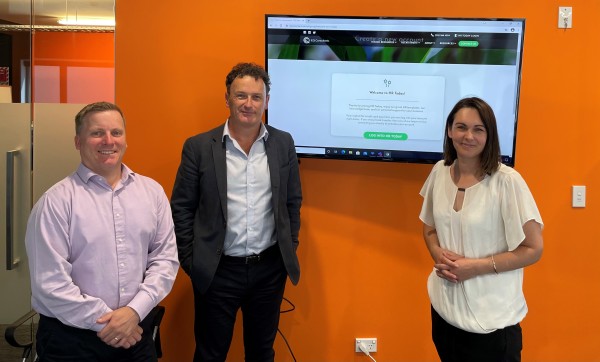 Two businessmen and one businesswoman stand in front of an orange wall and a TV screen featuring the HR Today website.
